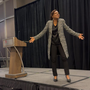 Dr. Regina Robinson, of the Massachusetts Department of Elementary and Secondary Education (DESE) standing on a stage, beside a podium, with her arms open, addressing the crowd.